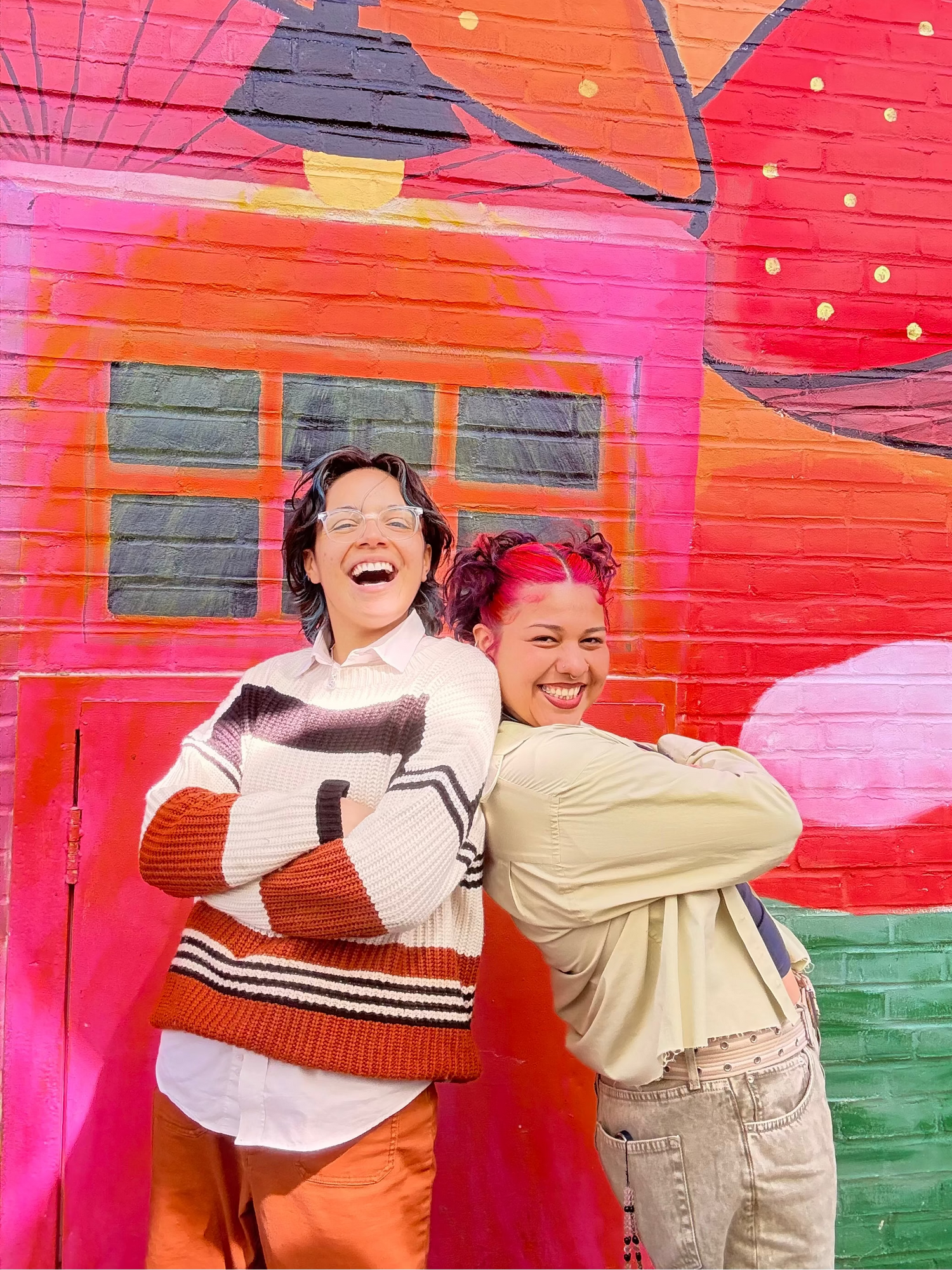 Two people posing in front of a colorful mural with a pink house and abstract shapes.